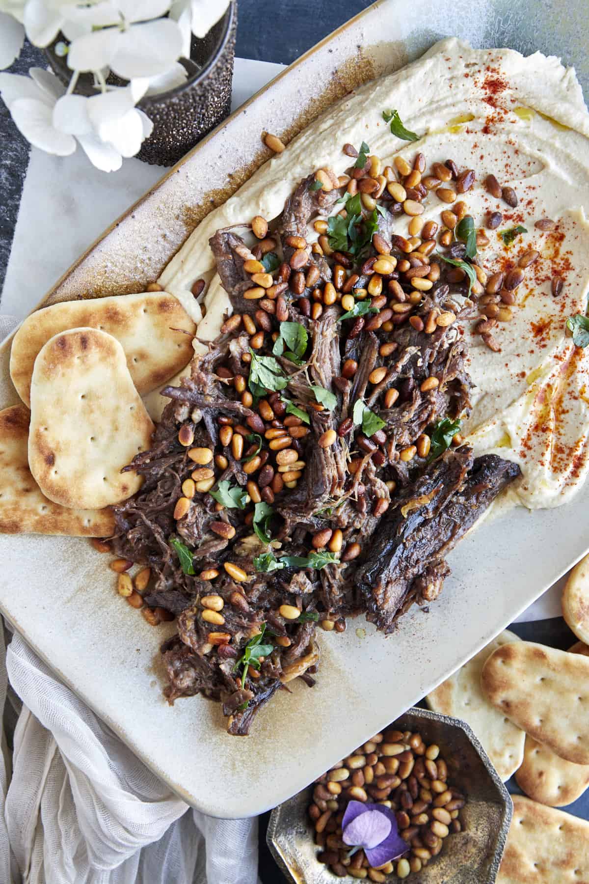 Hummus with meat on a platter topped with pine nuts and parsley with pita bread on the side. 