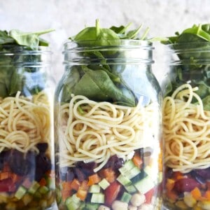 Three mason jar salads with chickpeas, veggies, spaghetti, spinach, and an olive oil dressing.