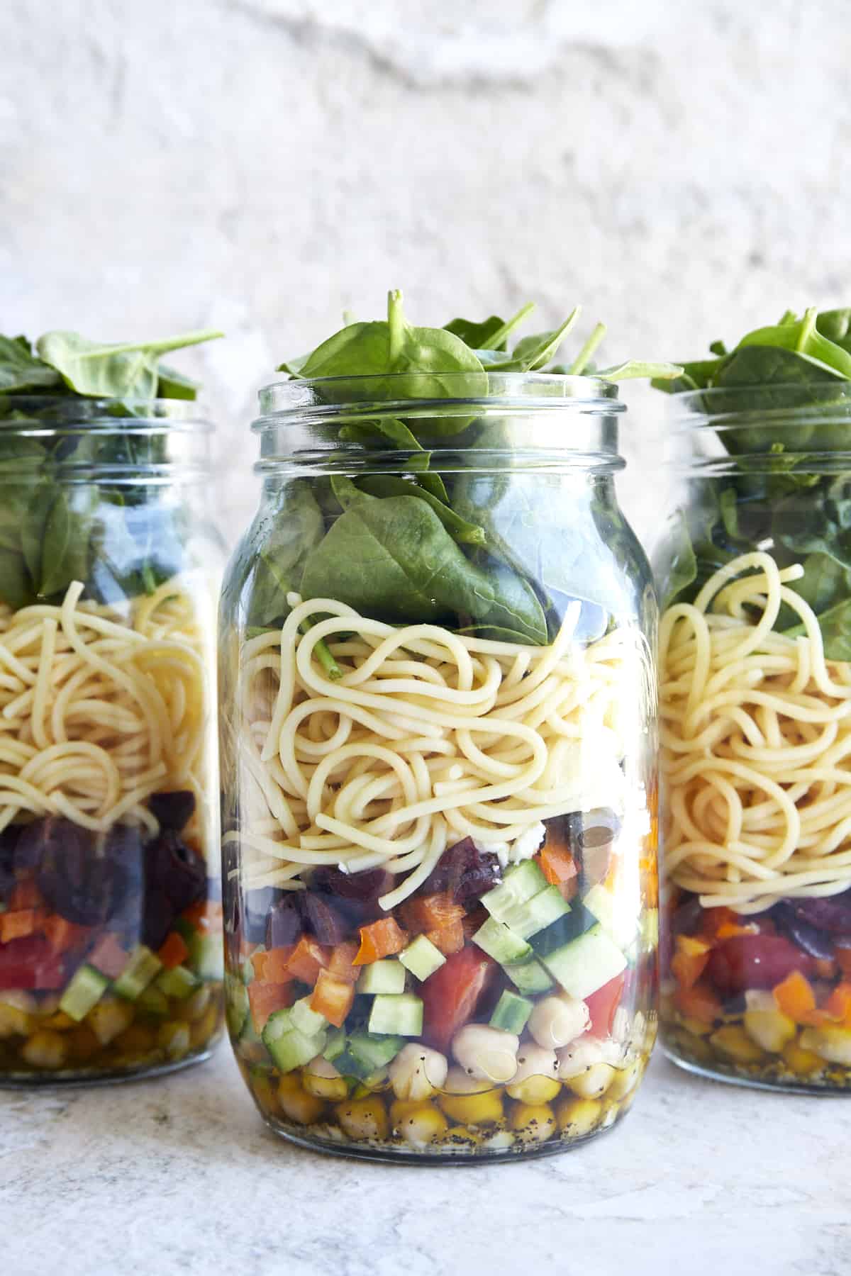 Three mason jar salads with chickpeas, veggies, spaghetti, spinach, and an olive oil dressing.