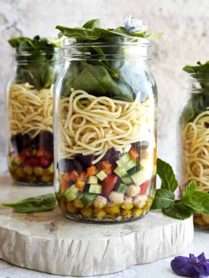 Three mason jar salads with an olive oil dressing in the bottom, chickpeas, chopped veggies, spaghetti, and spinach.
