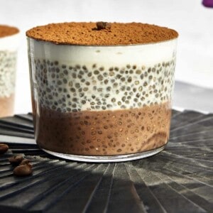 A jar of layered tiramisu chia pudding with a coffee layer, vanilla layer, yogurt layer, and cocoa powder on top.
