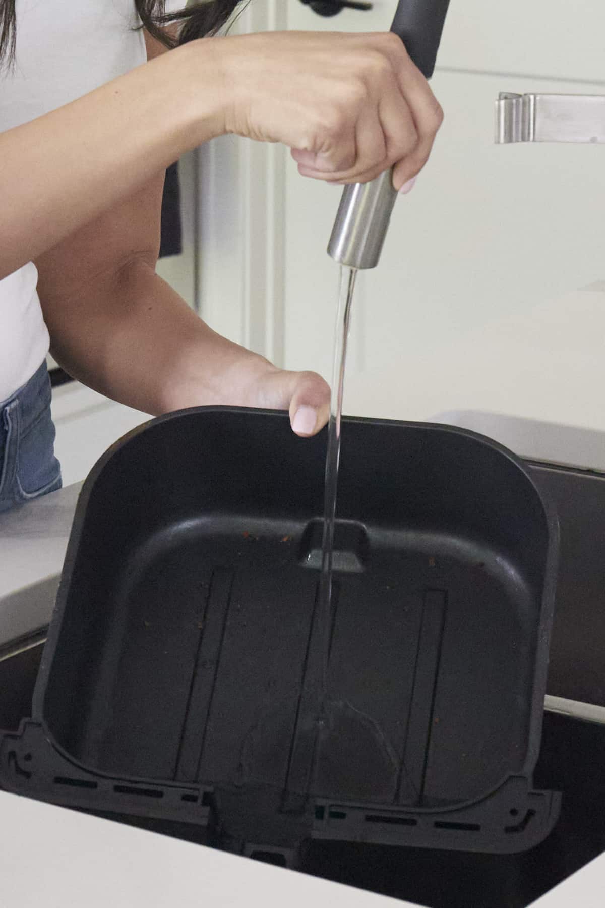 An air fryer basket being rinsed out with running water from a faucet in a sink. 