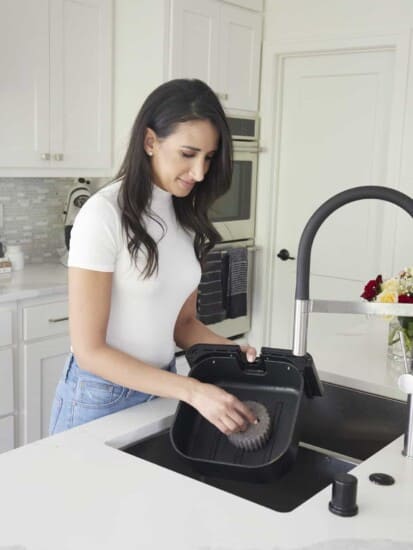 A woman using a sponge to clean the basket of an air fryer in a sink.