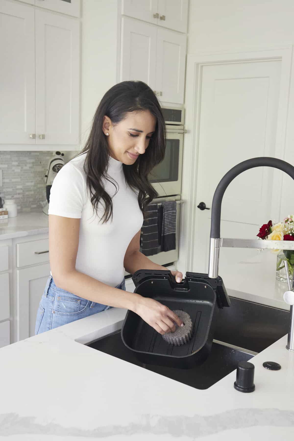A woman using a Scrub Daddy brush to clean the basket of an air fryer ina. sink. 
