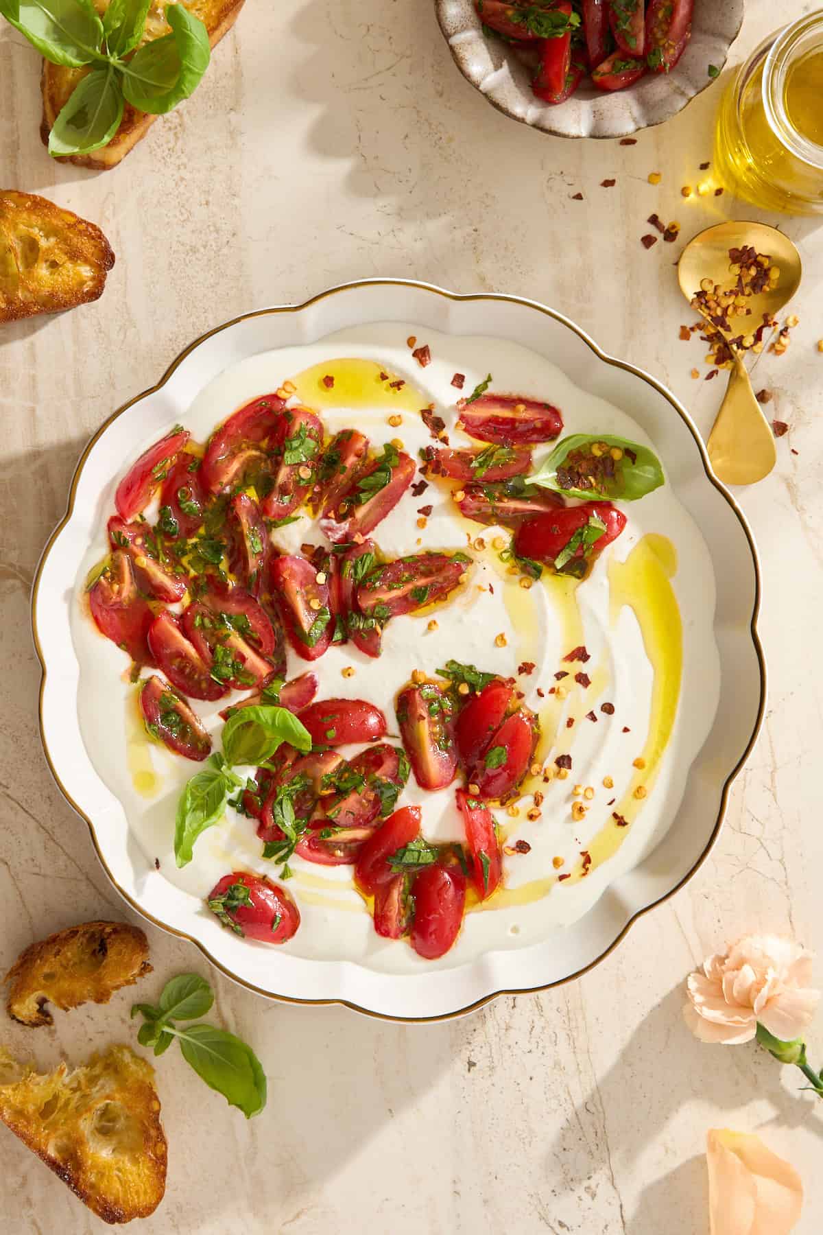 A white scalloped plate with sliced cherry tomatoes, fresh basil, olive oil, and red pepper flakes sits beside toasted bread and a bowl of tomatoes, while whipped cottage cheese adds creamy richness to this fresh dish on a light marble surface.