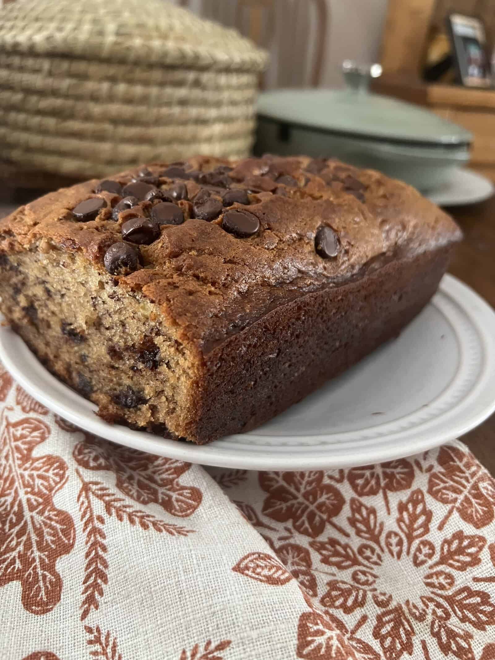 A loaf of chocolate chip banana bread sits on a white plate, showing a golden brown crust and chocolate chips on top. The plate is on a patterned cloth with leaves, and baskets are visible in the background.