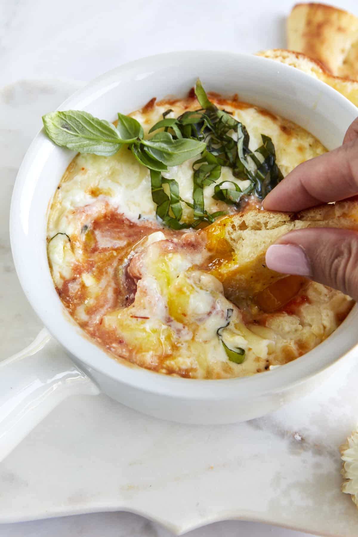 Two fingers dipping a piece of bread into a ramekin full of Eggs in Purgatory. 