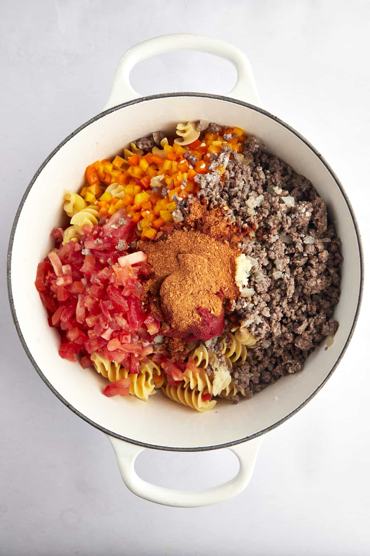 Overhead image of cooked beef, bell peppers, tomatoes, raw noodles, and taco seasonings in a pot. 