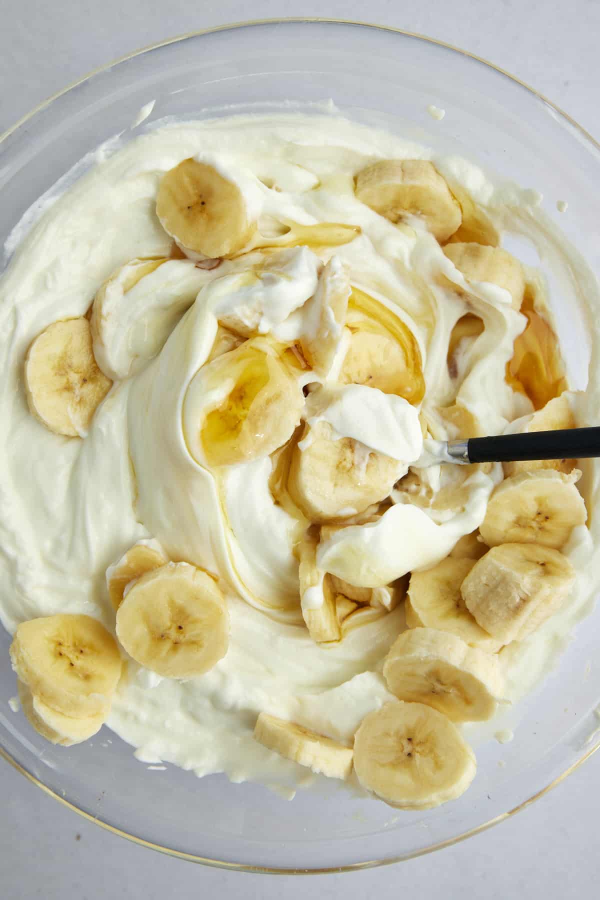 A mixture of Greek yogurt, honey, and banana coins in a mixing bowl with a spoon sticking out. 