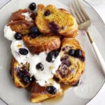 A plate full of croissant blueberry French toast topped with yogurt, blueberries, and maple syrup.