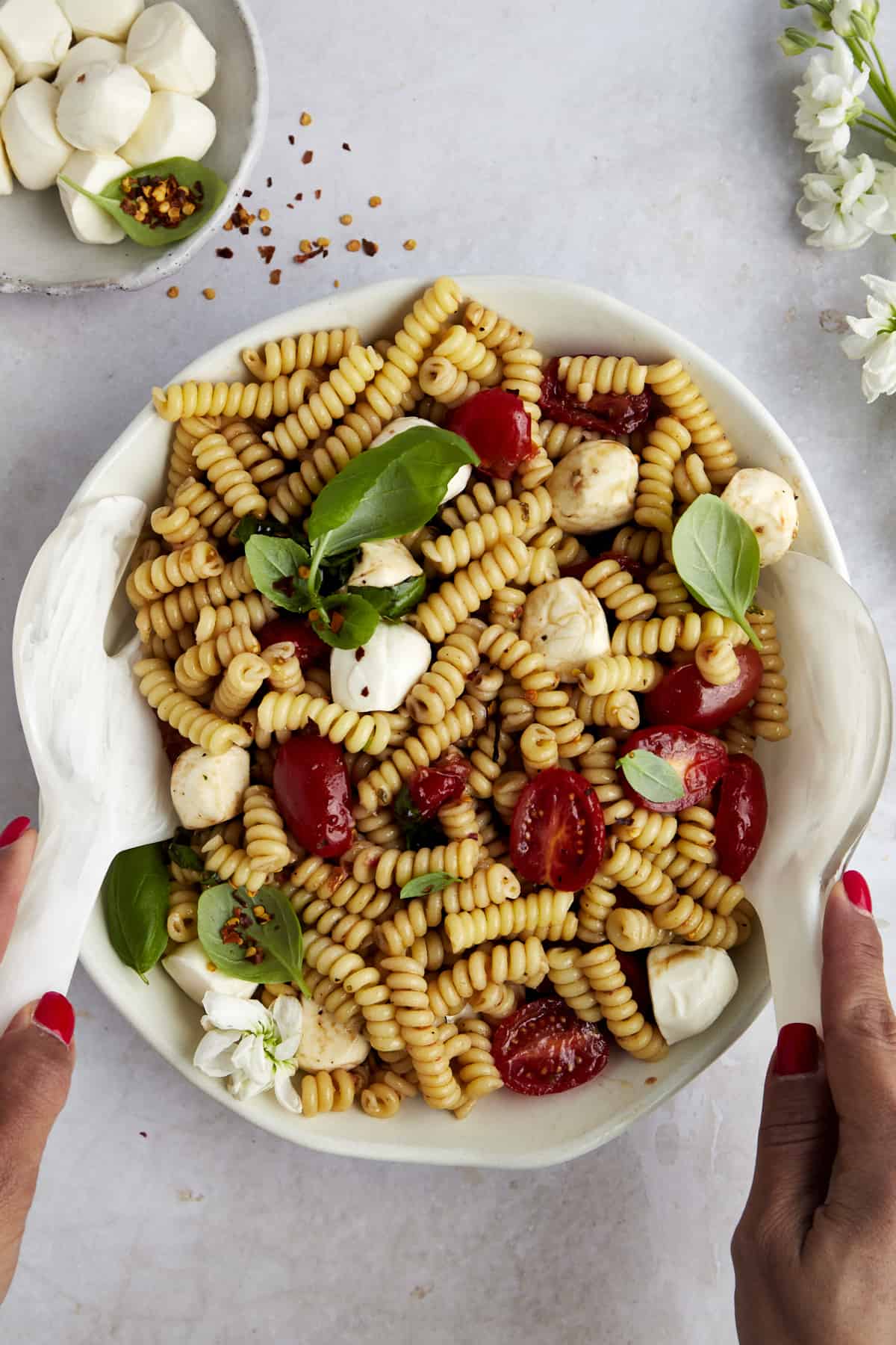 Two hands tossing caprese pasta salad in a bowl. 