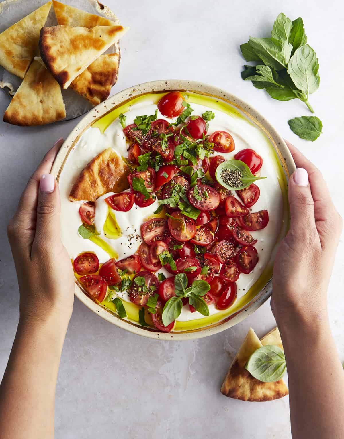 Two hands holding a bowl of marinated tomatoes and whipped feta dip with pita on the side.