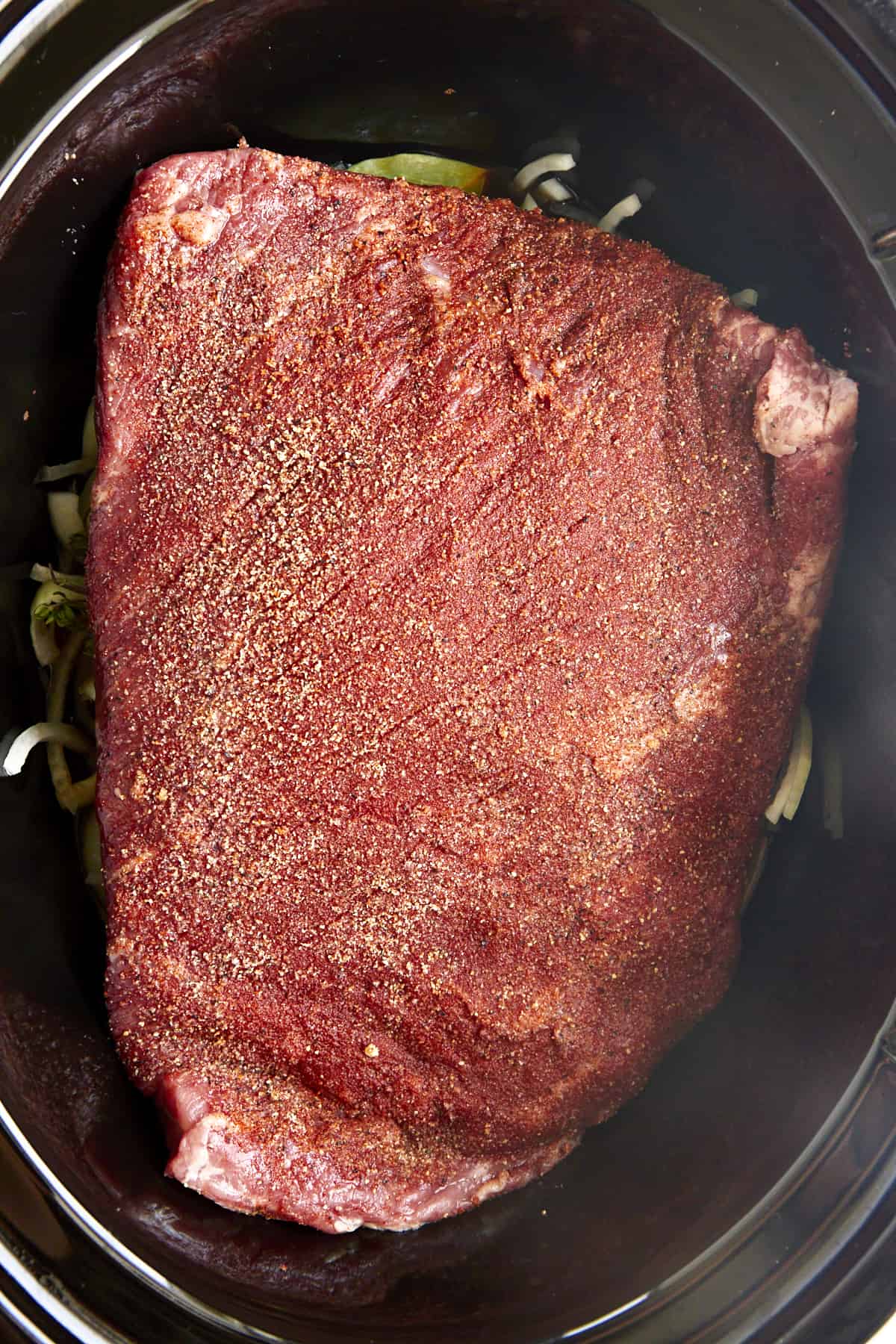 A 3-pound raw, seasoned beef brisket in a slow cooker with onions and herbs.