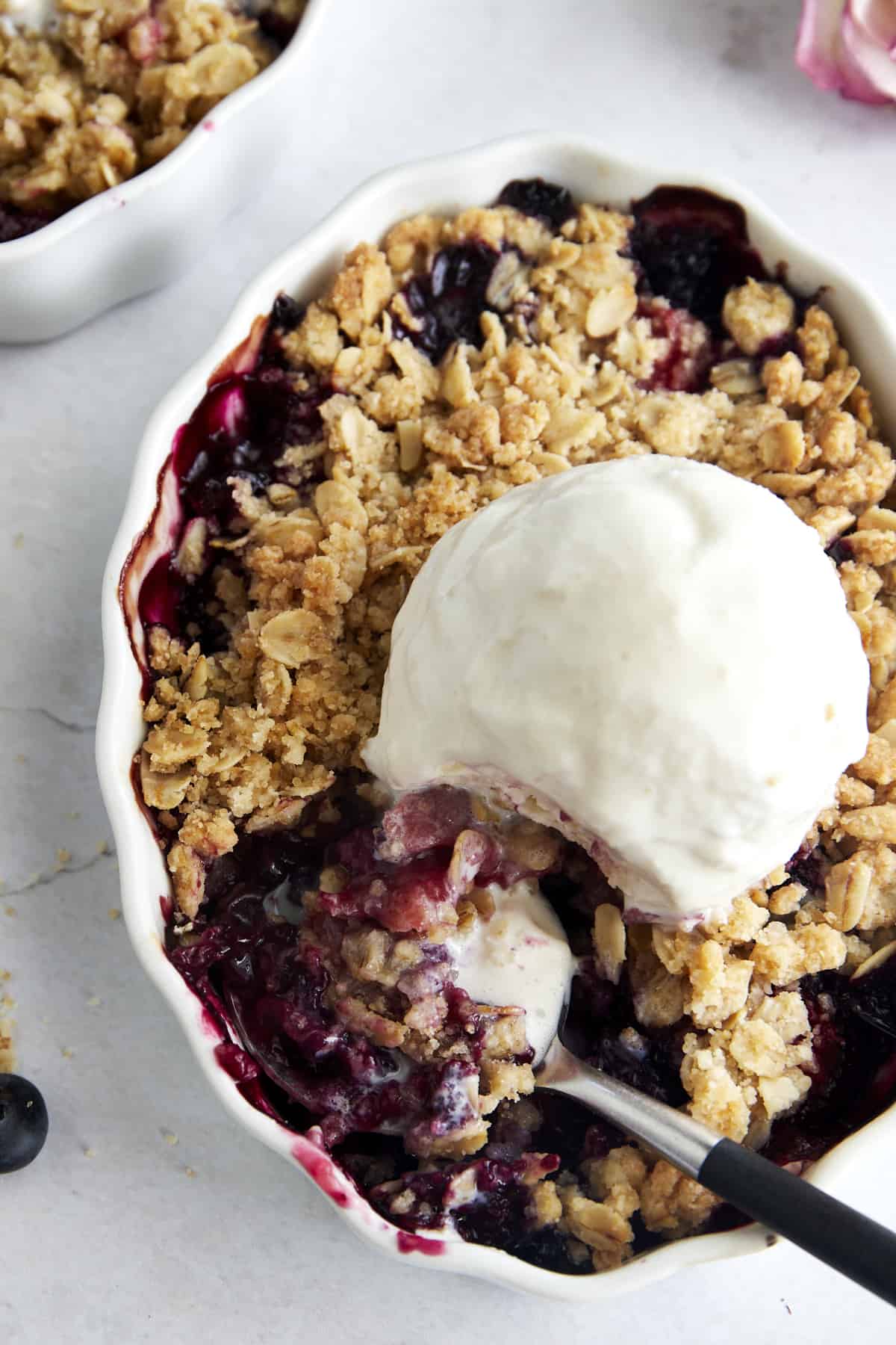 An oval baking dish full of mixed berry crisp topped with ice cream with a spoon taking a scoop.
