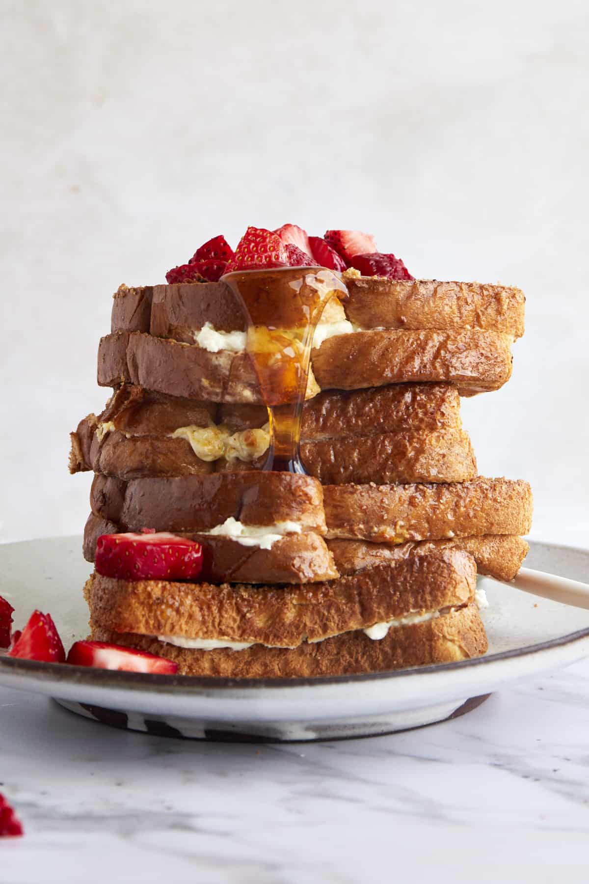 Air fryer french toast stuffed with cream cheese stacked on a plate topped with strawberries and syrup. 