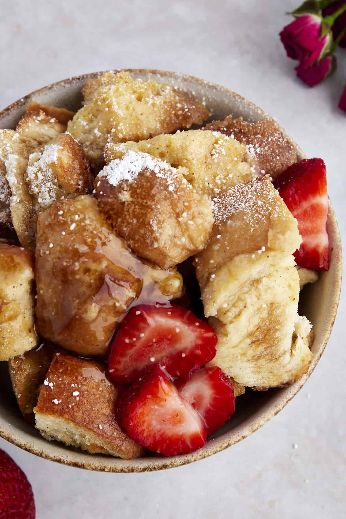 A bowl of crockpot French toast casserole topped with sliced strawberries, powdered sugar, and maple syrup. 
