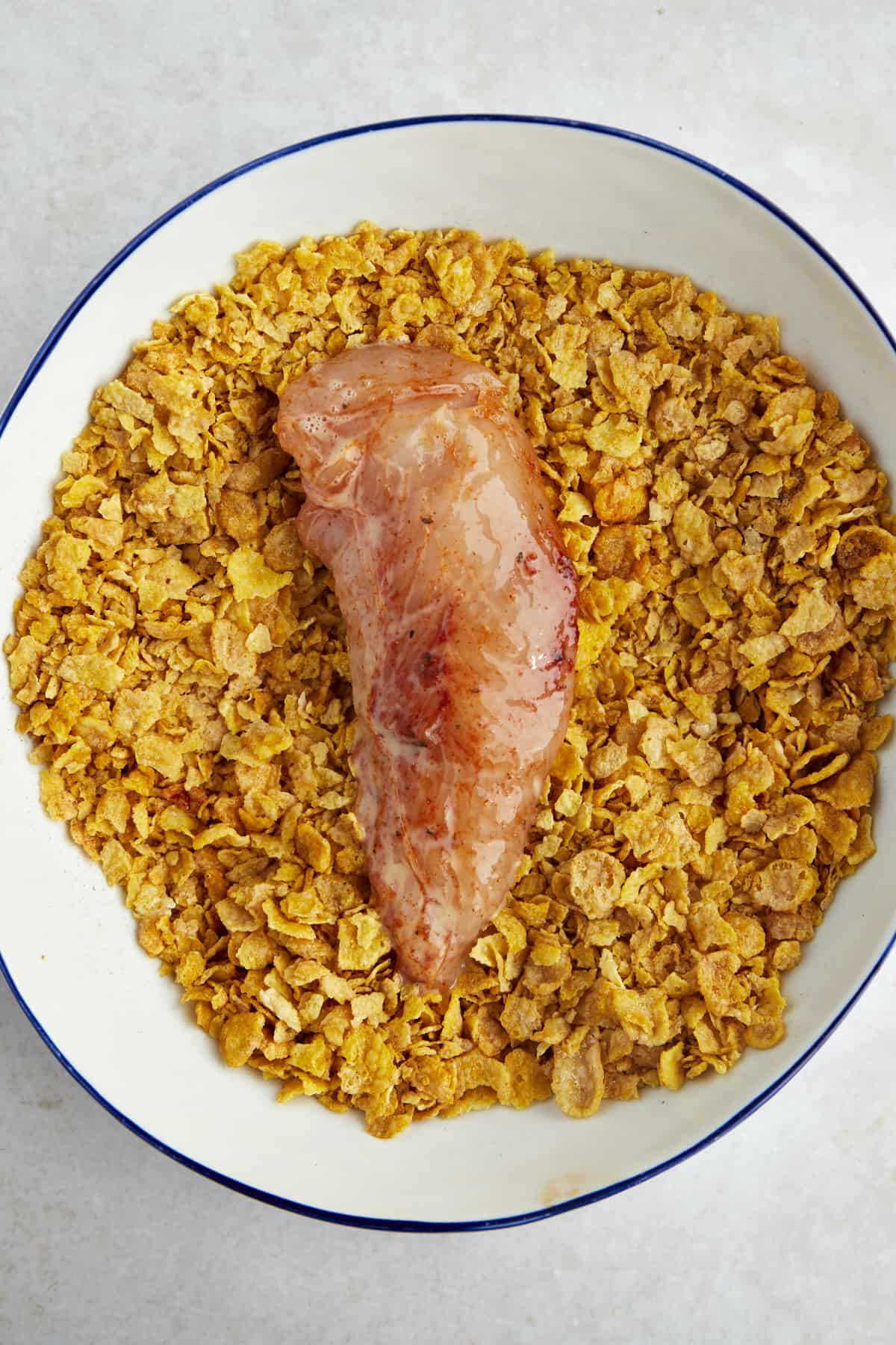 Raw chicken coated with eggs wash in a bowl of crushed cornflakes.