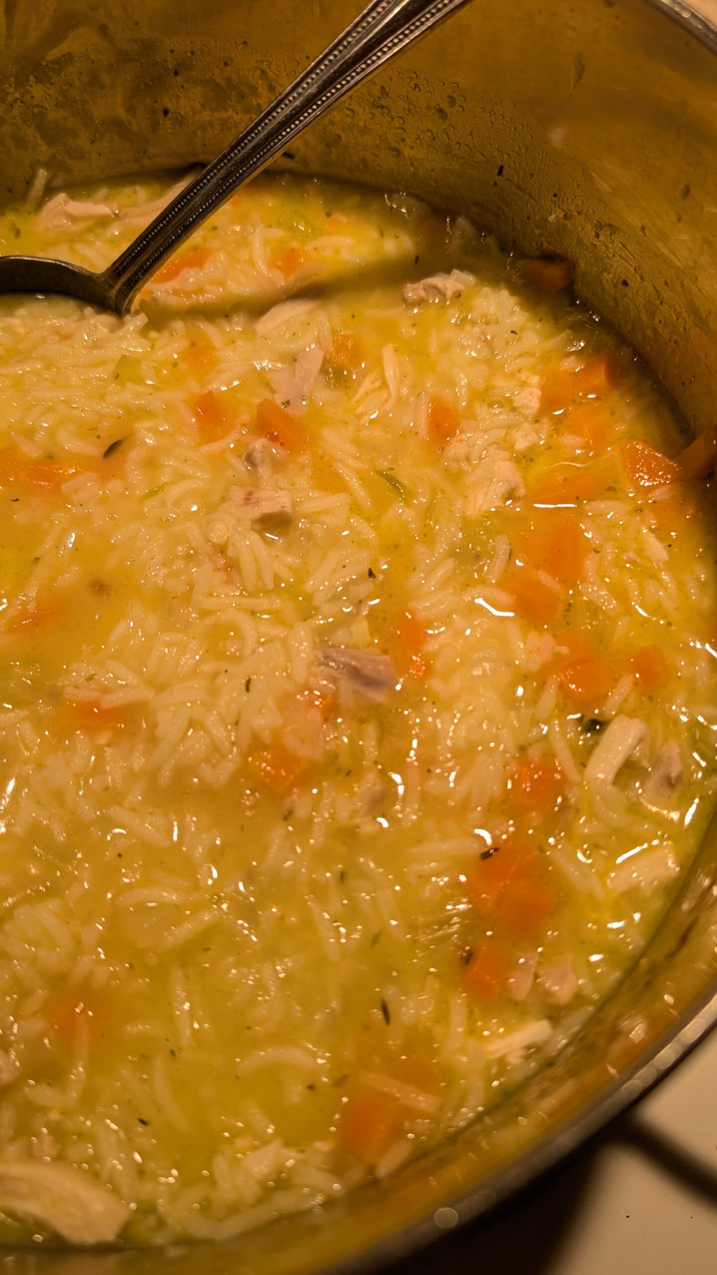 A close-up of a pot filled with chicken and rice soup, showing pieces of chicken, diced carrots, and rice in a yellowish broth. A black ladle rests on the rim of the pot.