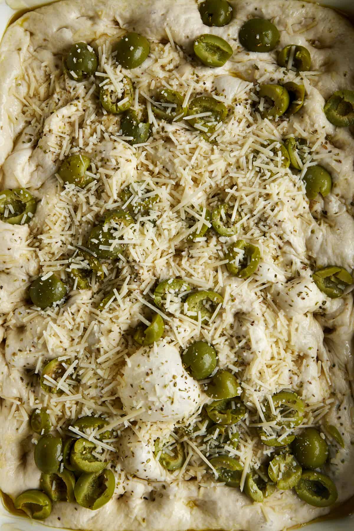 Raw focaccia dough topped with salt, oregano, rosemary, olives, and cheese.
