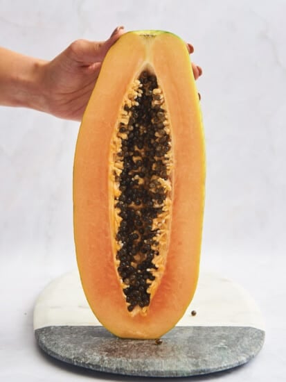 A halved papaya being held upright on a cutting board.