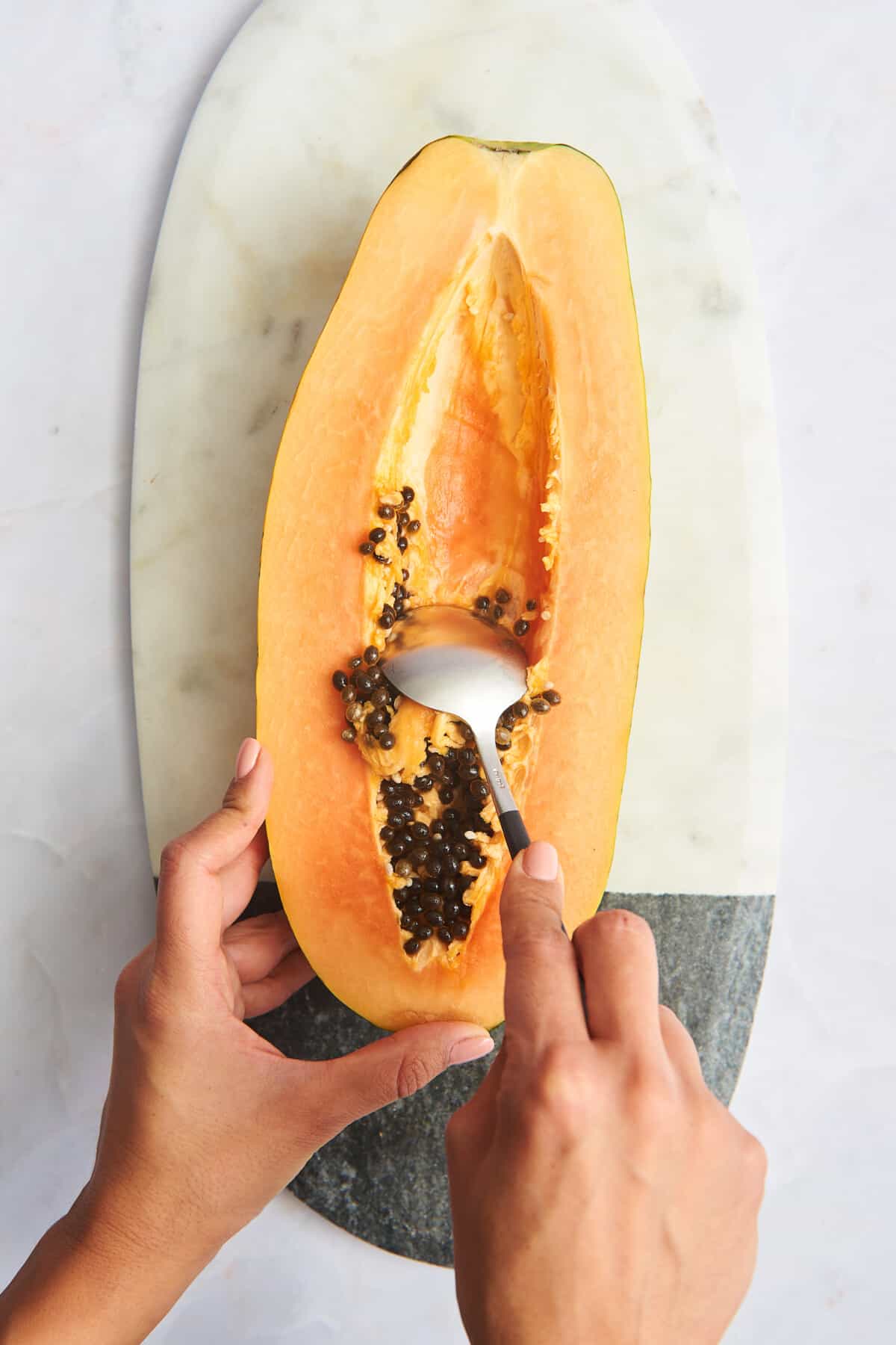 Two hands scooping out the black seeds of a papaya. 