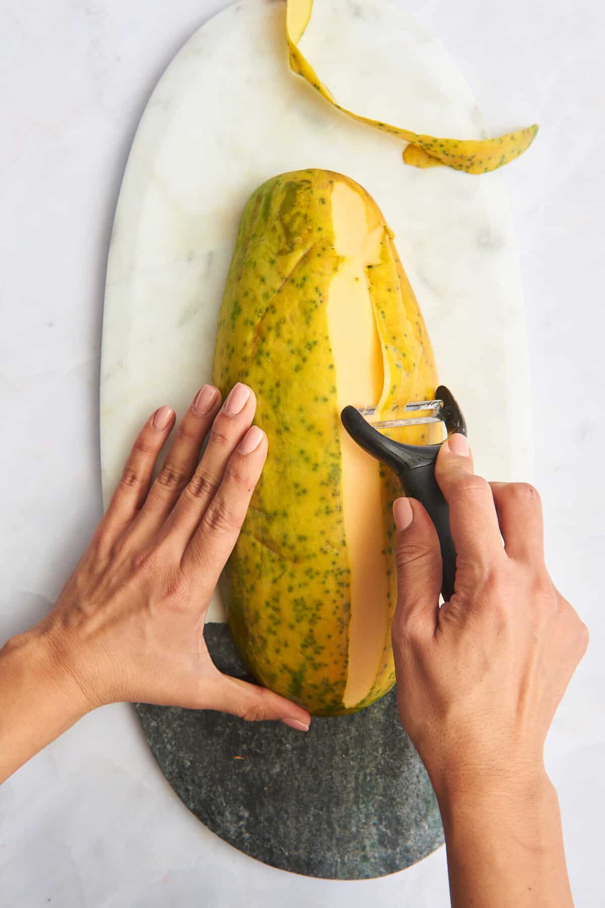A hand using a vegetable peeler to remove the skin from a papaya. 