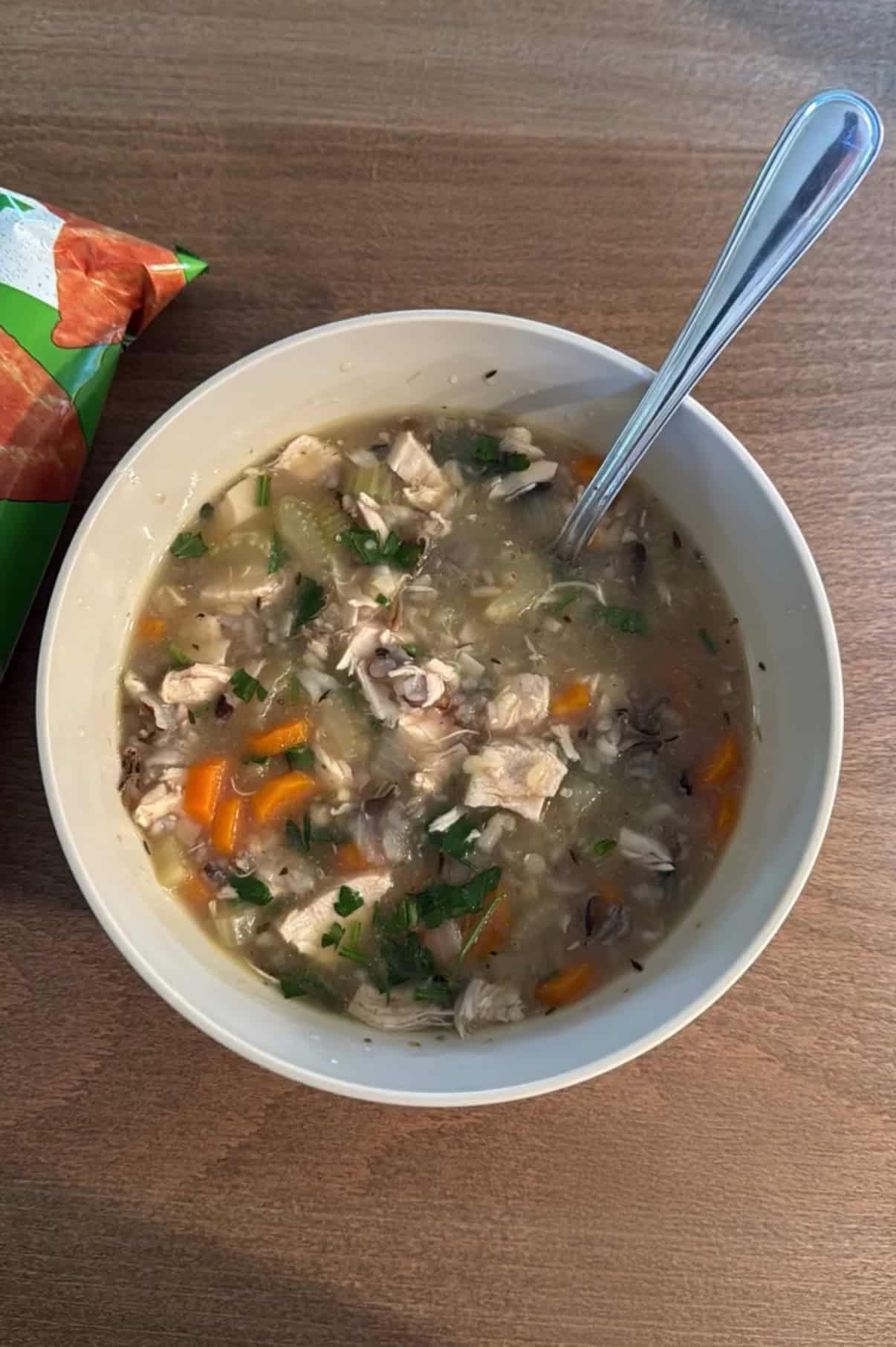 A bowl of homemade chicken and vegetable soup with pieces of chicken, carrots, celery, herbs, and rice, on a wooden table with a spoon in the bowl. A partially visible snack bag is beside the bowl.