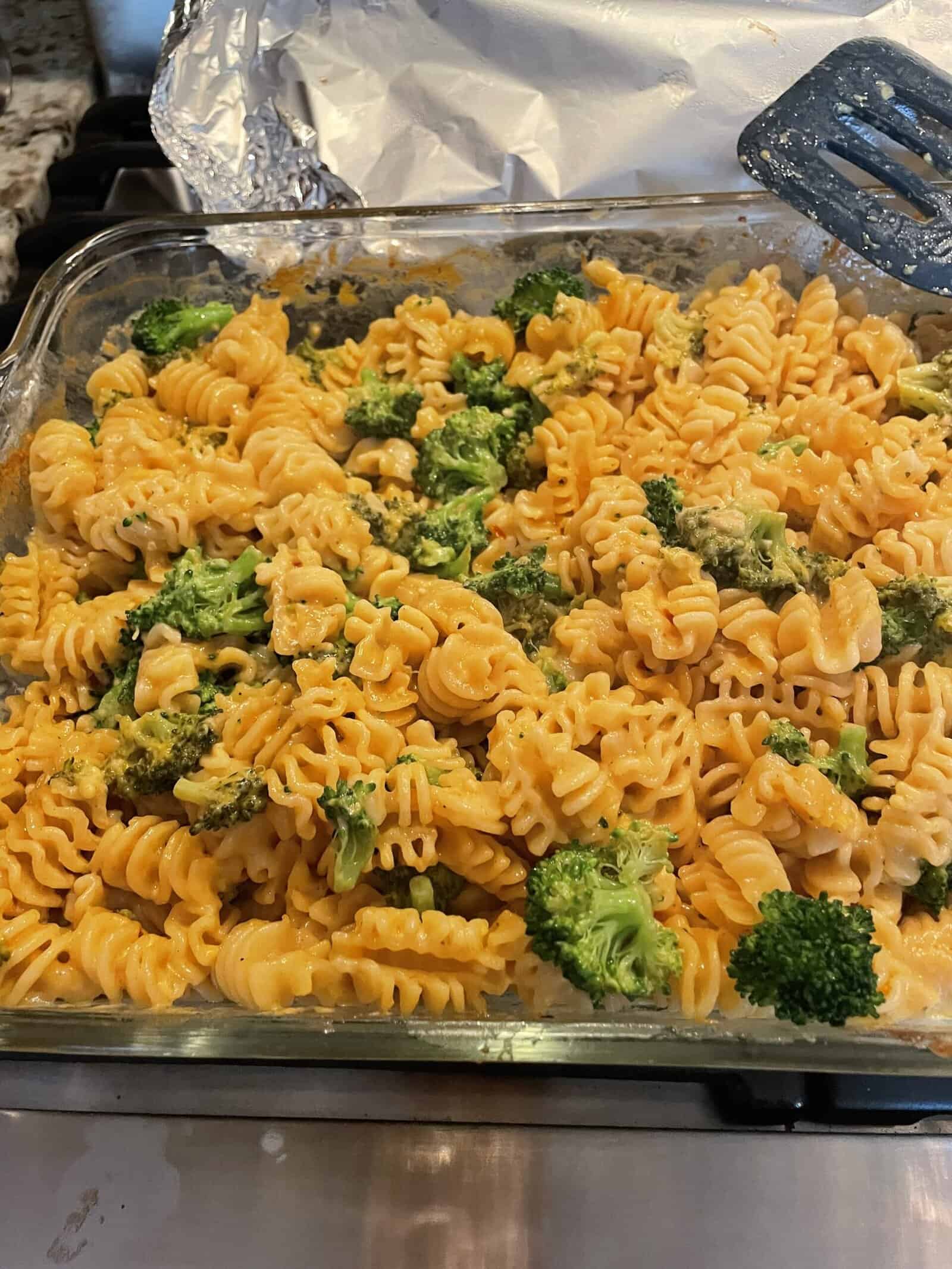 A glass baking dish filled with rotini pasta and broccoli, covered in melted cheese sauce, sits on a stovetop. A black spatula rests in the dish, and aluminum foil is in the background.