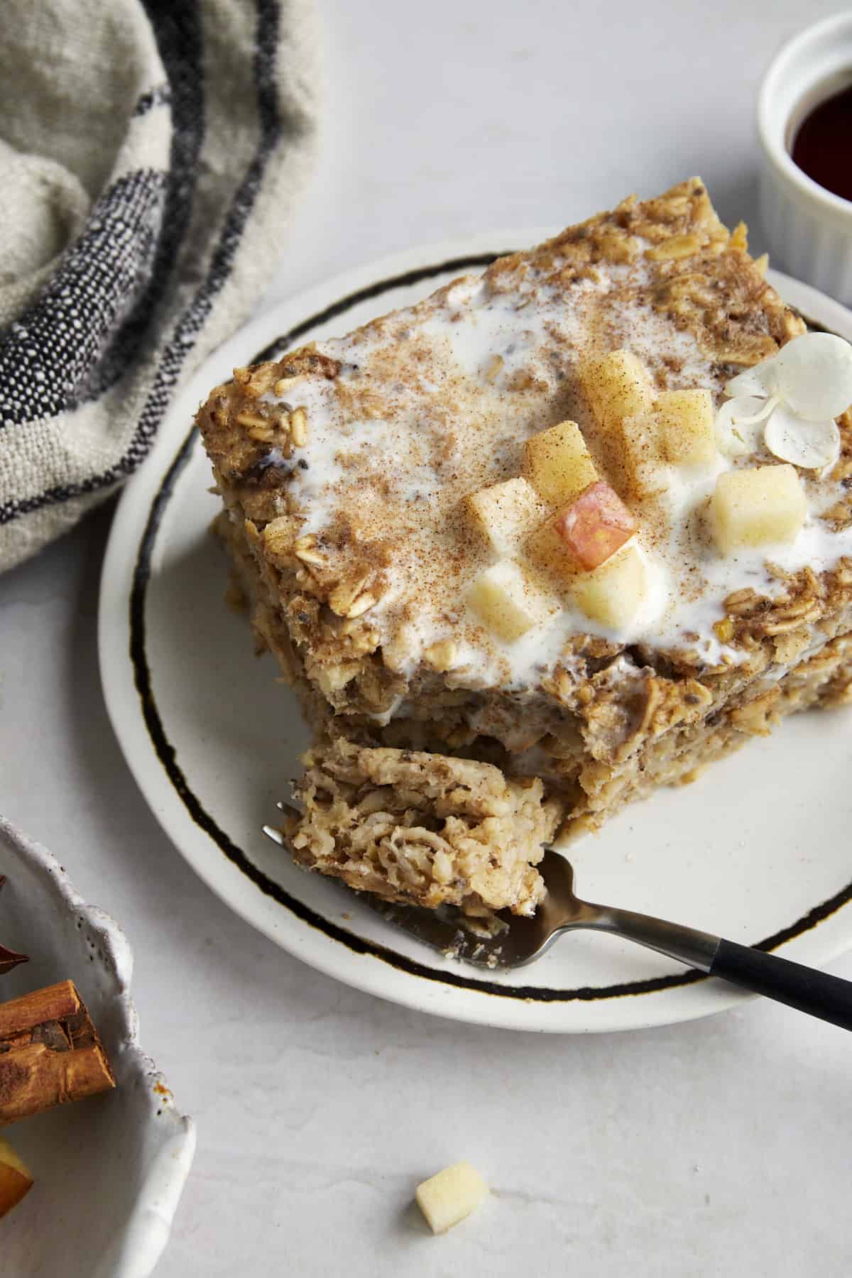 A slice of baked apple oatmeal on a plate topped with apples and milk with a fork holding a bite. 