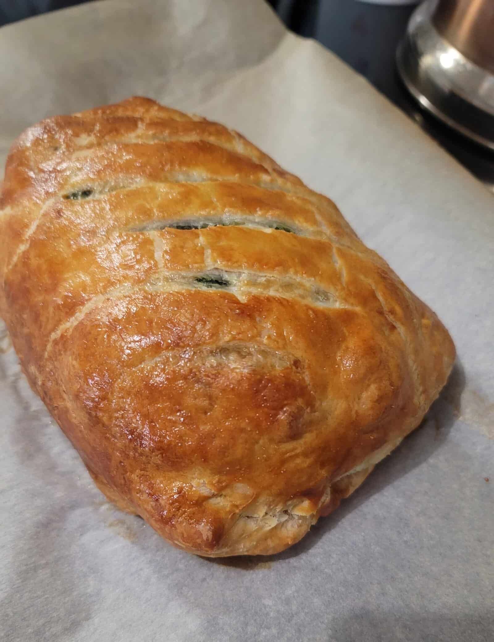 A golden-brown, flaky pastry loaf with diagonal slits on top, resting on a sheet of parchment paper.