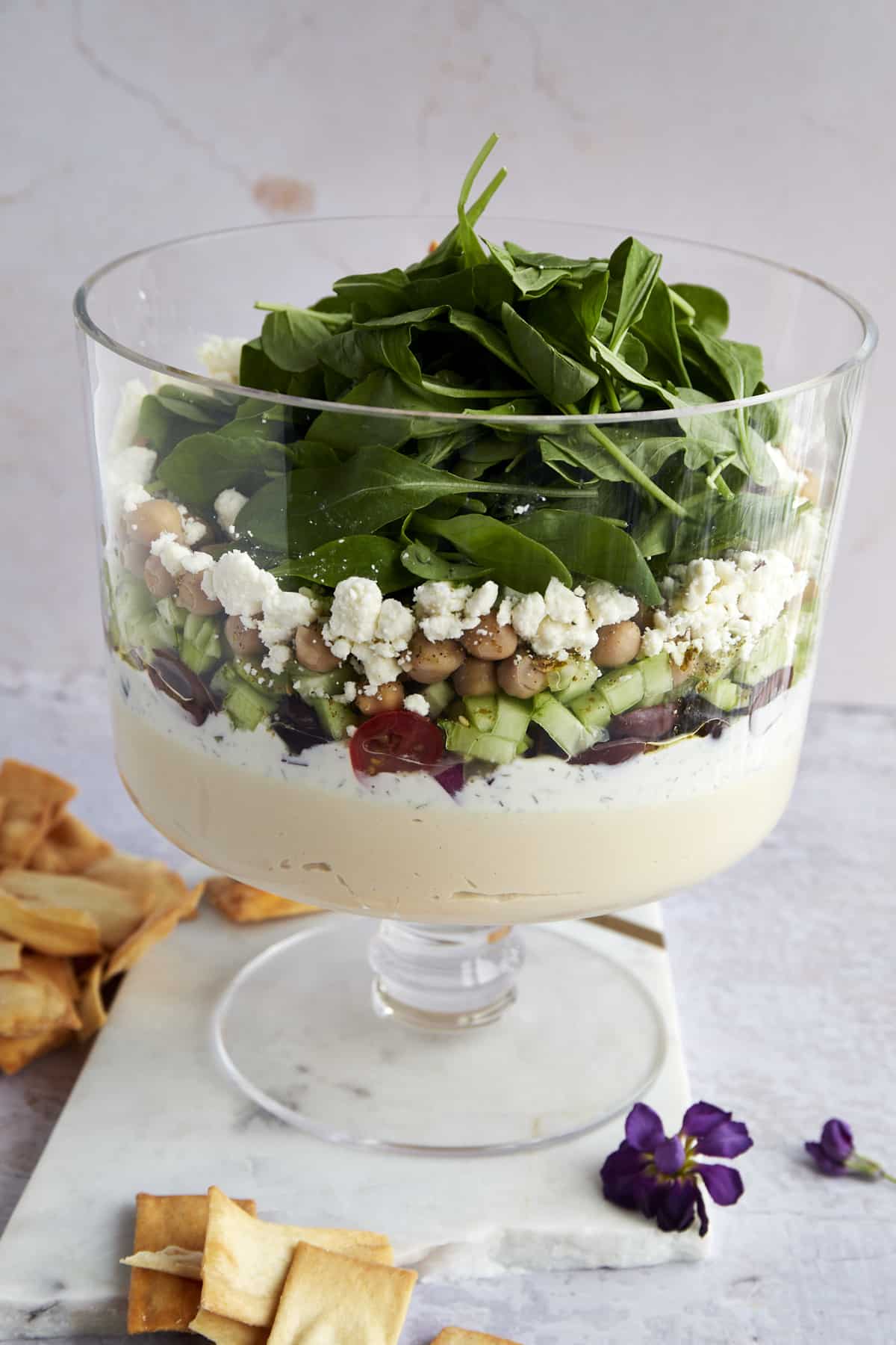 Greek dip with hummus, tzatziki, tomatoes, cucumbers, olives, feta, and arugula in a trifle bowl. 
