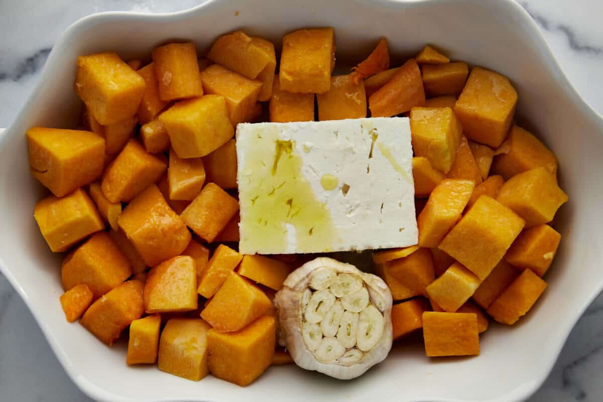 Butternut squash cubes, a block of feta, and a head of garlic topped with oil in a baking dish. 
