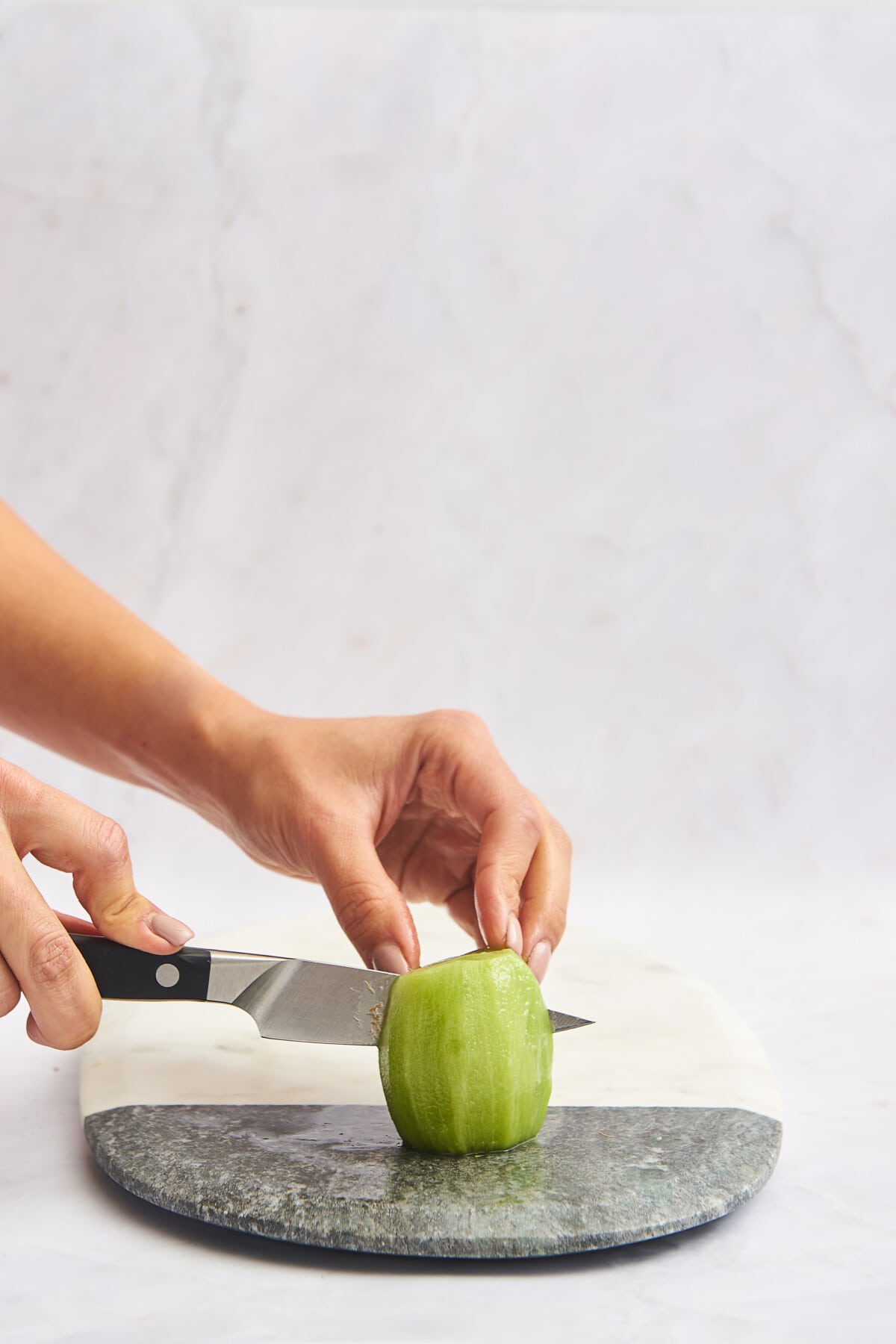 A hand using a knife to slice a skinless kiwi in half. 