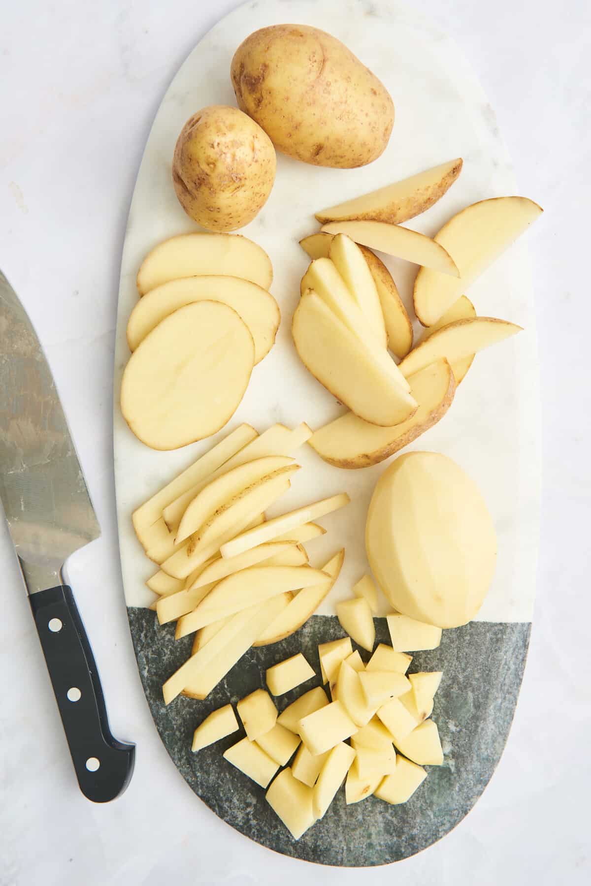 Whole potatoes, peeled potatoes, potato slices, wedges, fries, and cubes on a cutting board. 