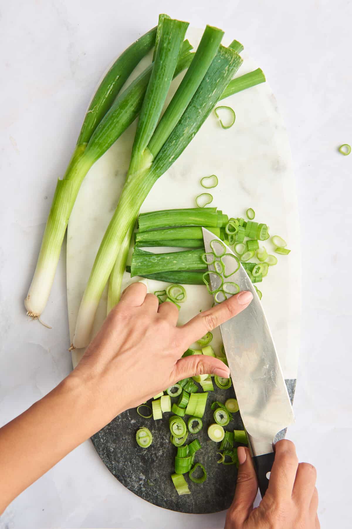A knife with chopped scallions over a cutting board with whole and chopped green onions. 