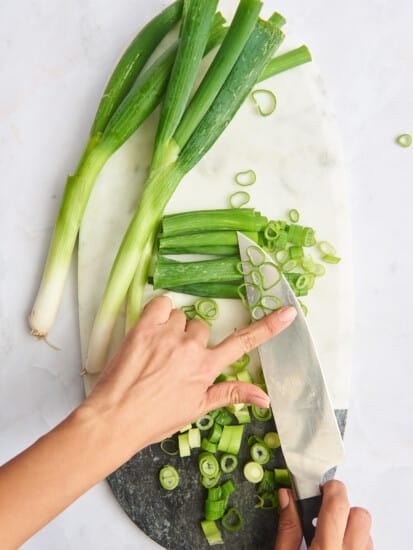 Scallions being slid off a knife onto a cutting board with whole and chopped scallions.