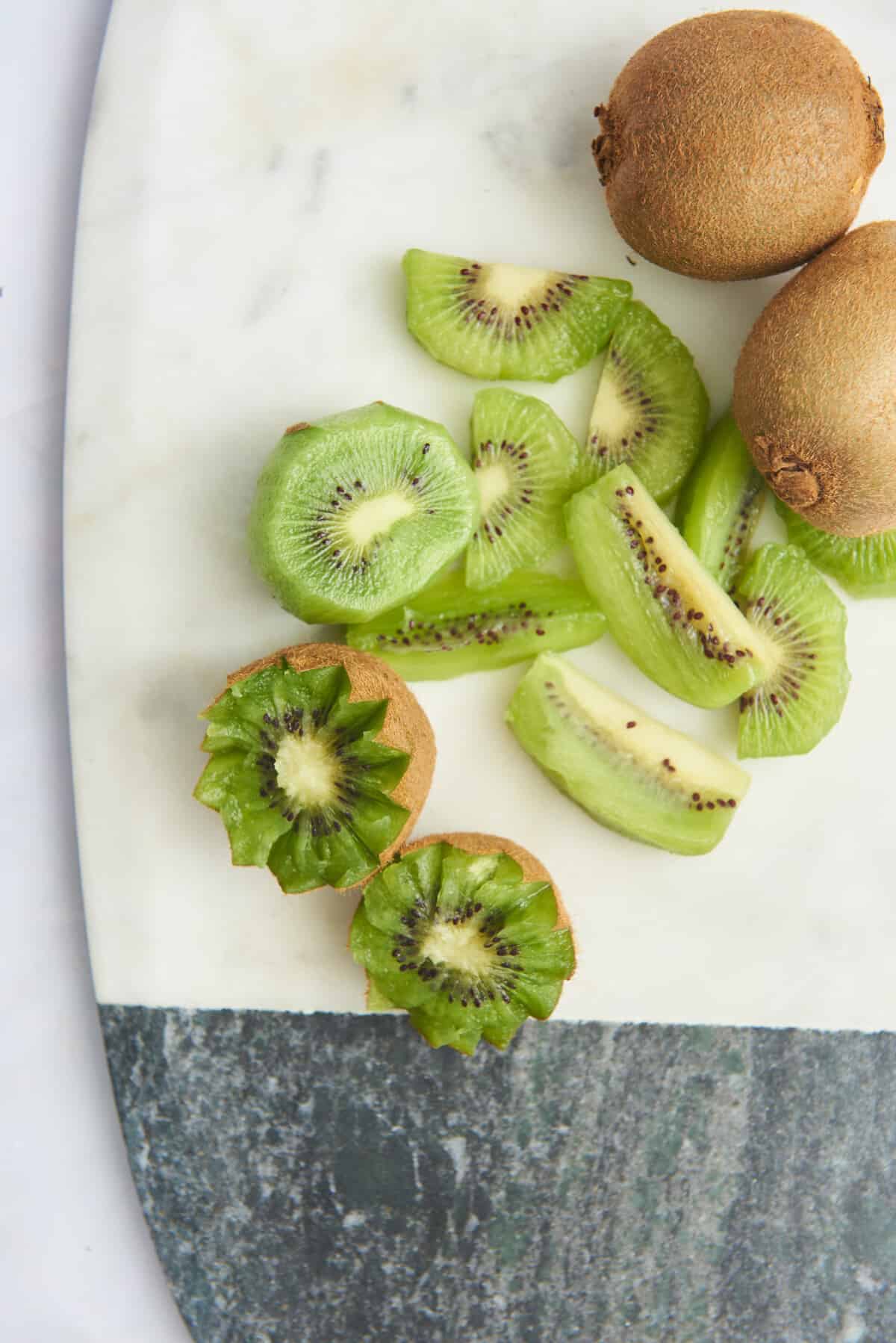 Whole kiwis, kiwi wedges, and star shaped kiwi on a marble cutting board. 