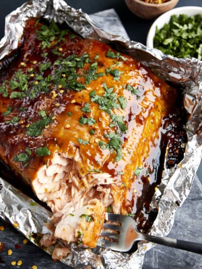 A fork tearing a bite of honey garlic salmon from a filet in a foil packet.