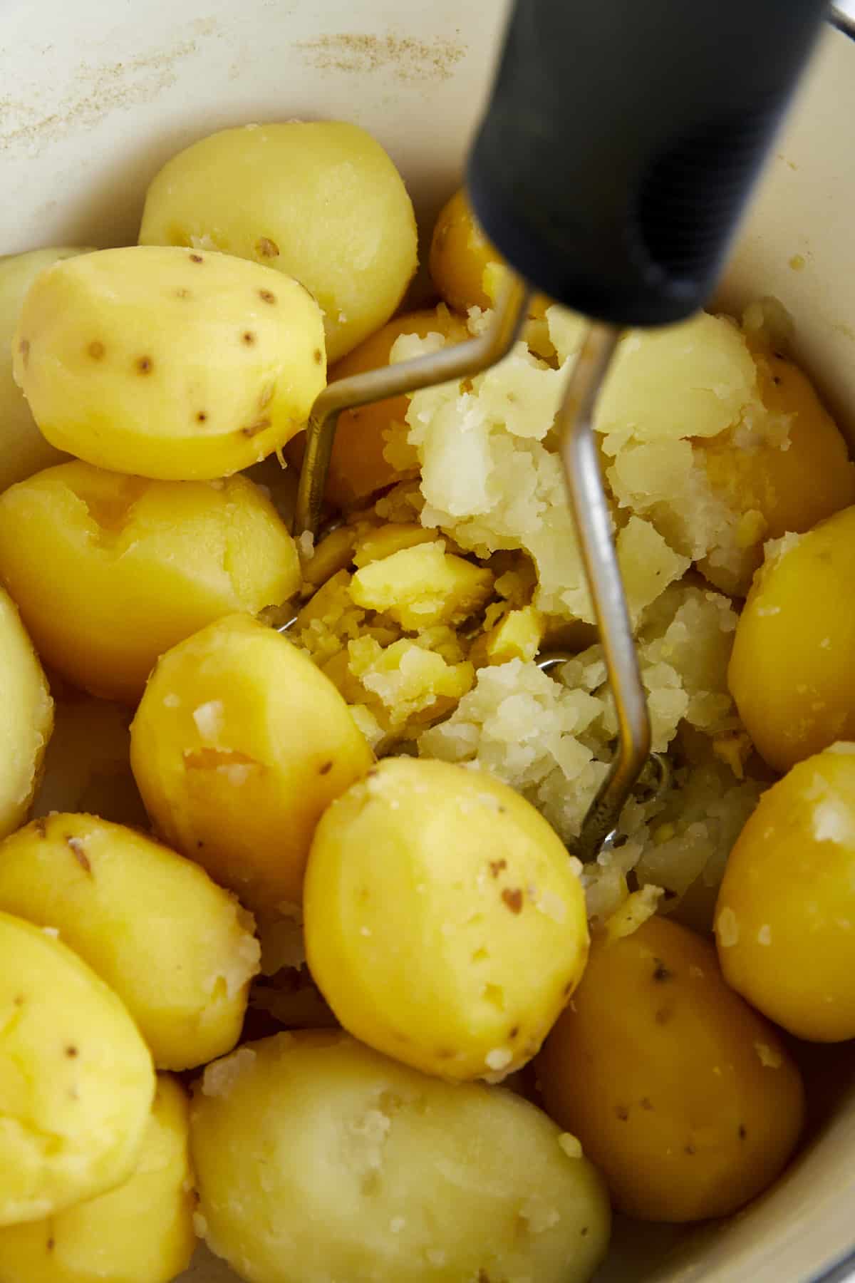 Boiled potatoes being mashed with a potato masher in a pot. 