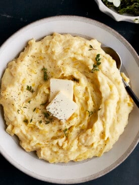 A bowl of the best mashed potatoes topped with butter, pepper, and herbs.