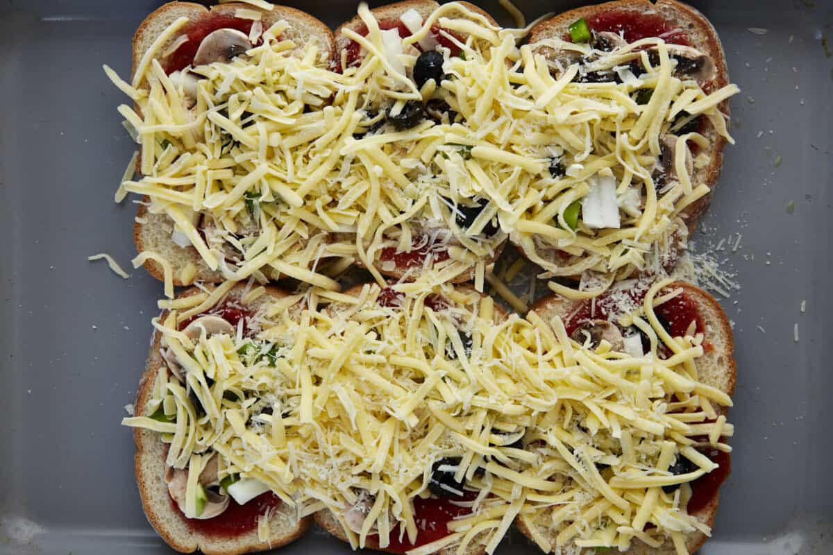 Shredded cheese sprinkled over slices of bread topped with marinara, peppers, onions, and mushrooms on a baking sheet. 