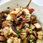 A spoon lifting a scoop of Mediterranean style stuffing from a baking dish.