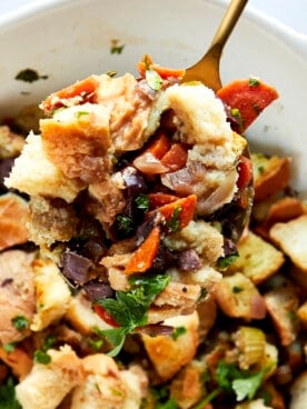 A spoon lifting a scoop of Mediterranean style stuffing from a baking dish.