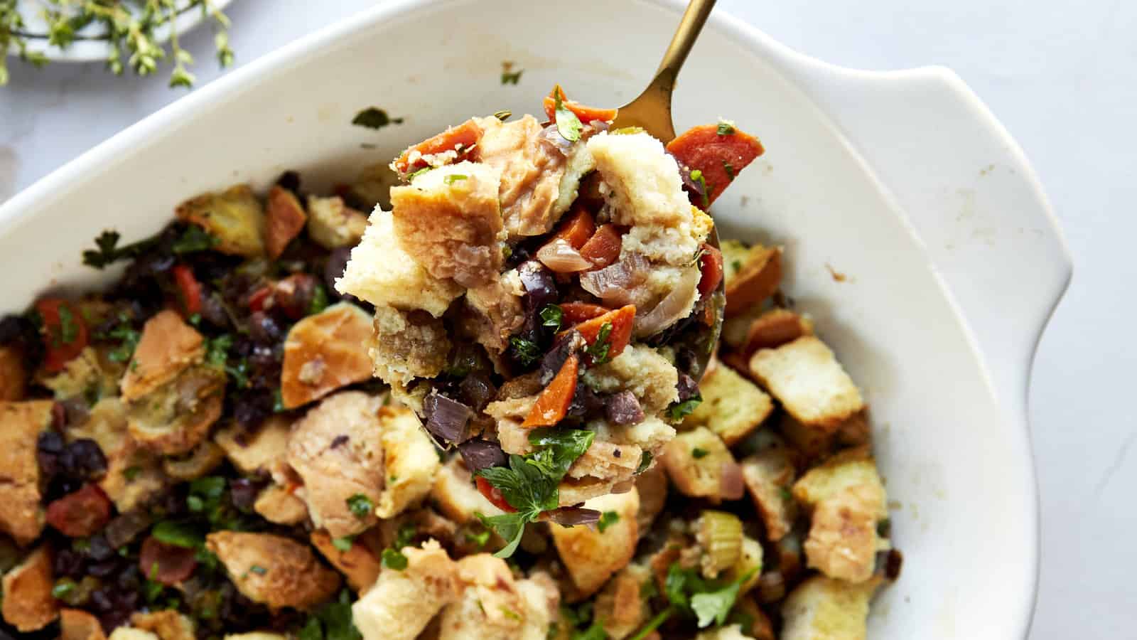 A spoon lifting a scoop of Mediterranean style stuffing from a baking dish.