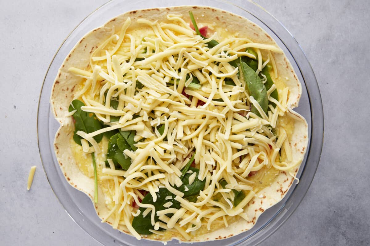 Cheese and veggies over an unbaked egg and cottage cheese mixture over a tortilla in a round dish. 