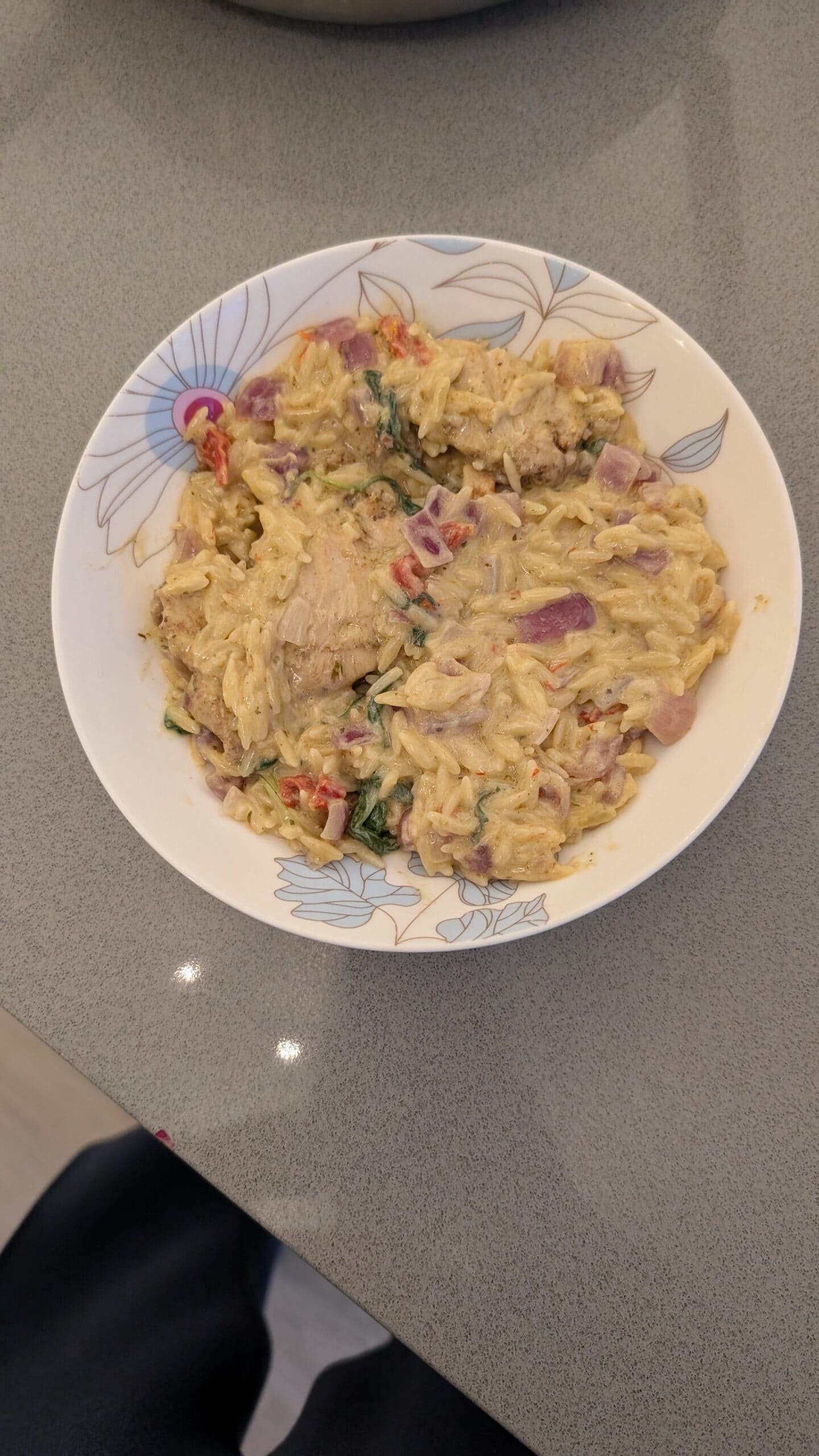 A bowl of creamy pasta with shredded chicken, red onions, and herbs, served in a white floral-patterned dish on a gray countertop.