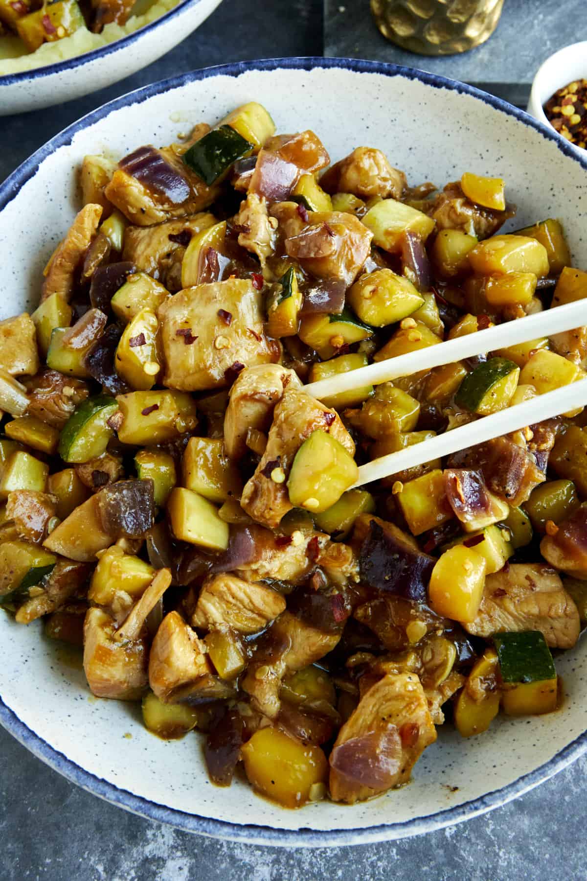 Two chopsticks picking up a bite of chicken and zucchini stir fry. 