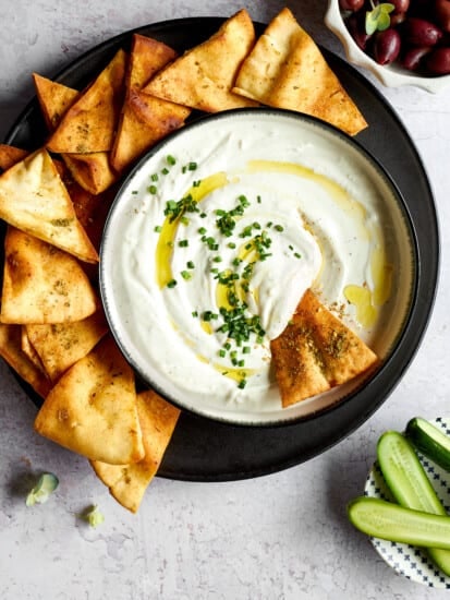 A pita chip dipped into a bowl of garlic yogurt sauce surrounded by more pita chips.