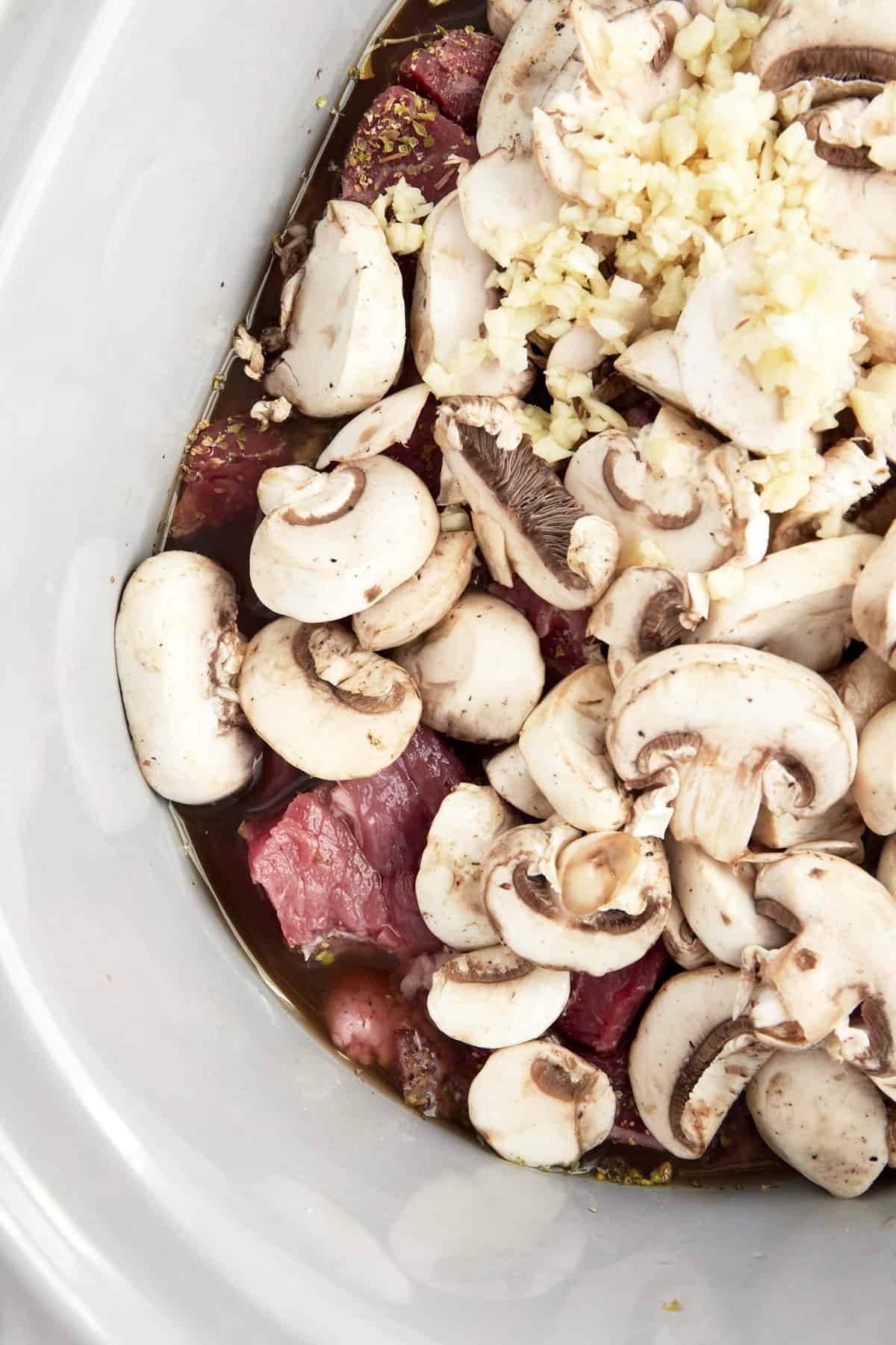 Uncooked beef, mushrooms, garlic, and sauce in a slow cooker. 