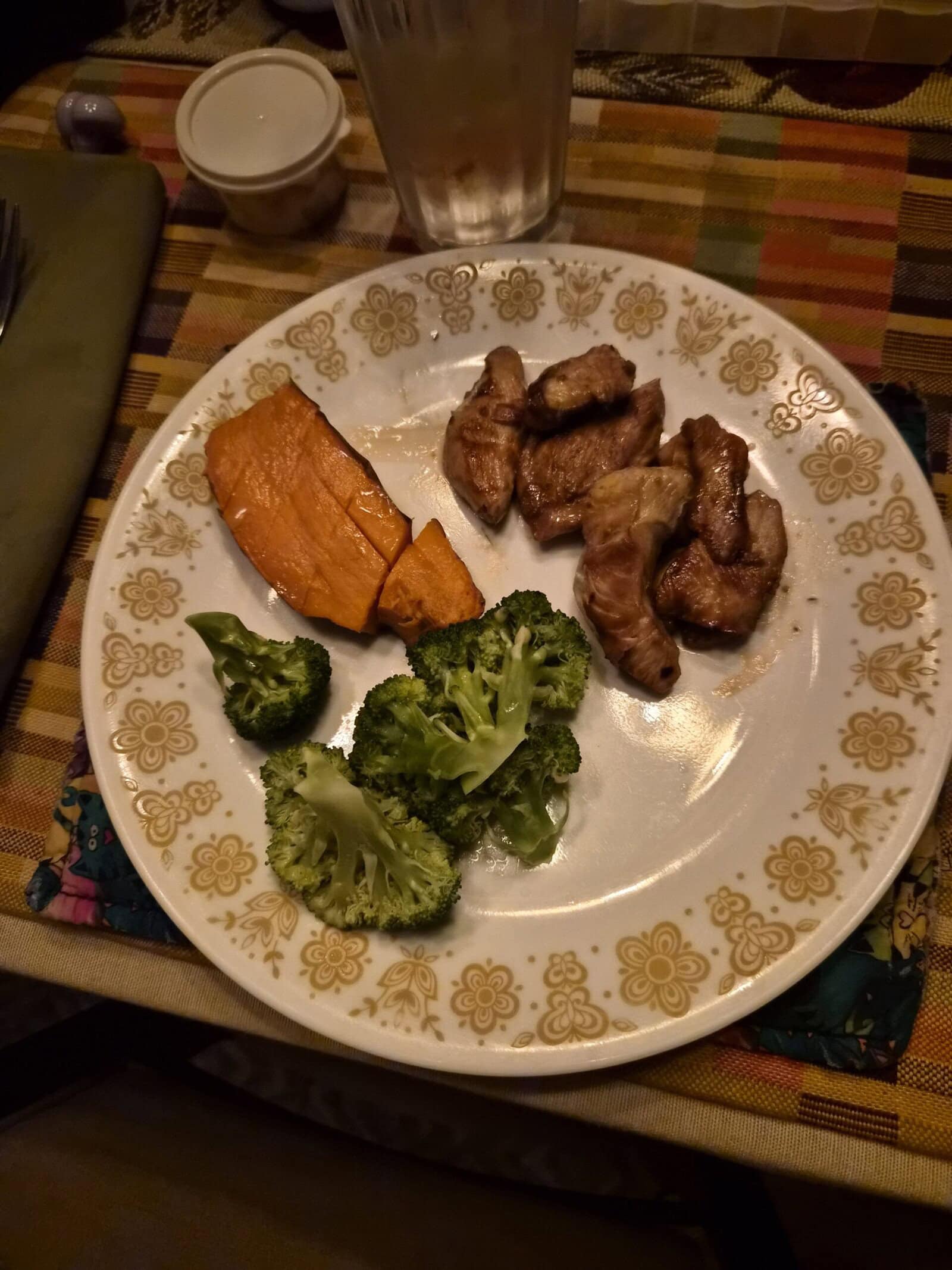 A plate with pieces of cooked meat, two broccoli florets, and a roasted sweet potato slice on a patterned tablecloth. A glass of water and small container are in the background.